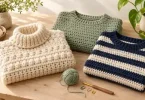 Three handmade crochet sweaters in cream, sage, and navy stripes with yarn and crochet tools